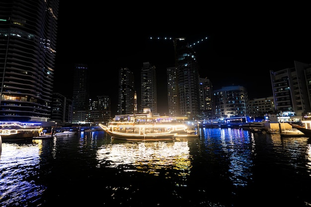 Небоскребы, освещенные ночью, отражаются в воде канала Дубай Марина Бэй с яхтами и гиперлапсом замедленной съемки лодок