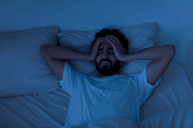 Slaapprobleem gestrest jonge Indiase man ligt in bed en raakt zijn hoofd aan