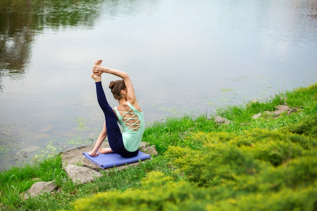 Slank Kaukasisch donkerbruin meisje dat yoga in de zomer op een groen gazon door de rivier doet