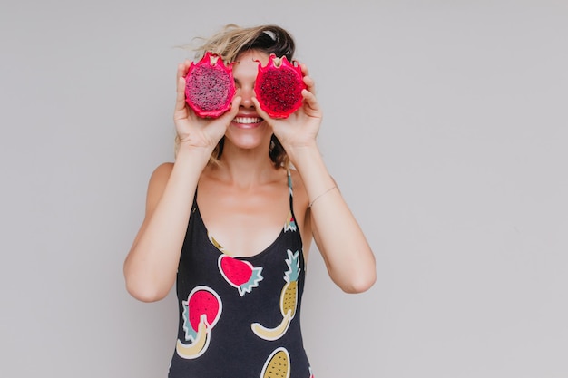 Slank meisje dat positieve emoties uitdrukt terwijl ze poseert met rode pitahaya Studioportret van blij vrouwelijk model dat plezier heeft tijdens fotoshoot met exotisch fruit