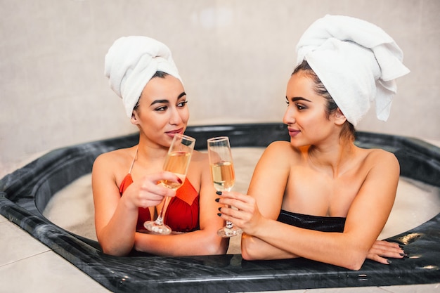 Slanke en sexy jonge vrouwen zitten in een hydromassagebad en kijken elkaar aan. Modellen raken glazen met champaigne. Hun lichaam is omwikkeld met witte handdoeken.