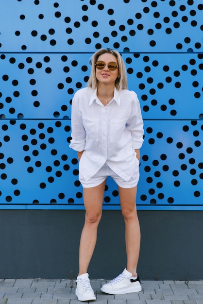 Slanke jonge blonde vrouw in een wit pak trekt grimassen en heeft plezier in de zomer tegen een blauwe muur