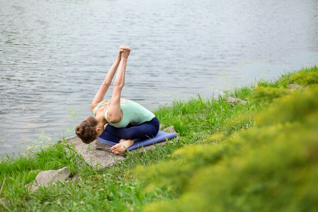 Slanke jonge brunette yoga voert uitdagende yoga-oefeningen op groen gras uit tegen een achtergrond van de natuur