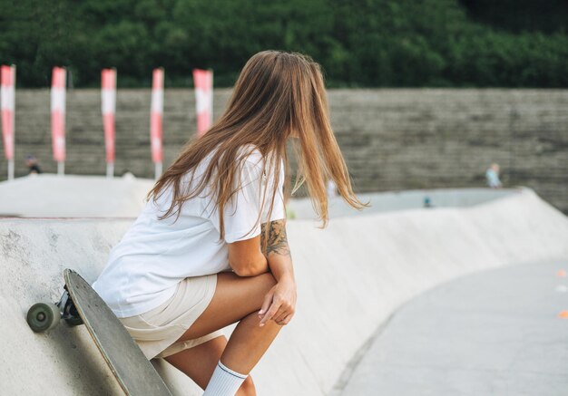 Slanke jonge vrouw met lang blond haar in lichte sportkleding met longboard in skatepark bij zonsondergang