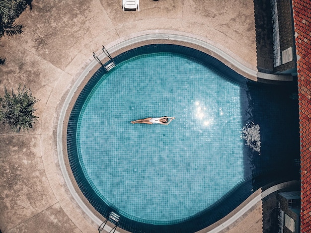 Slanke meditatieve dame drijft in het ronde blauwe zwembad van een tropisch hotel, haar armen omhoog, vlekjes in het schone water. Modeconcept, drone-weergave