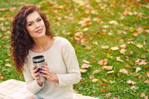 Slanke mooie jonge vrouw rusten in de herfst in het park en zittend op het gazon op het gazon en het drinken van thee uit een thermocup