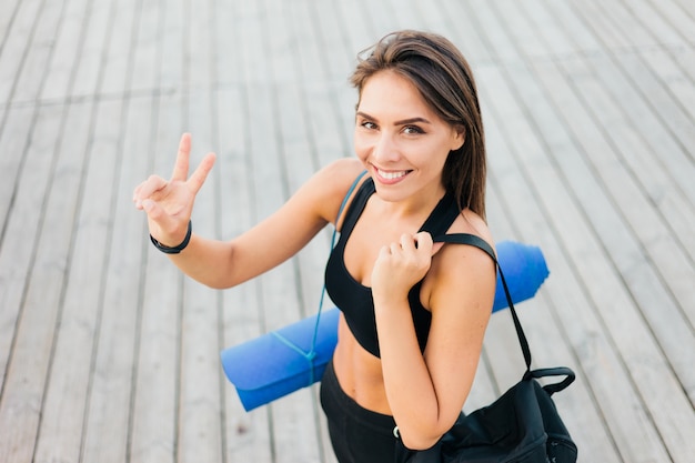 Slanke vrolijke fit vrouw in sportkleding met sporttas en mat op haar schouders poseren na training buitenshuis