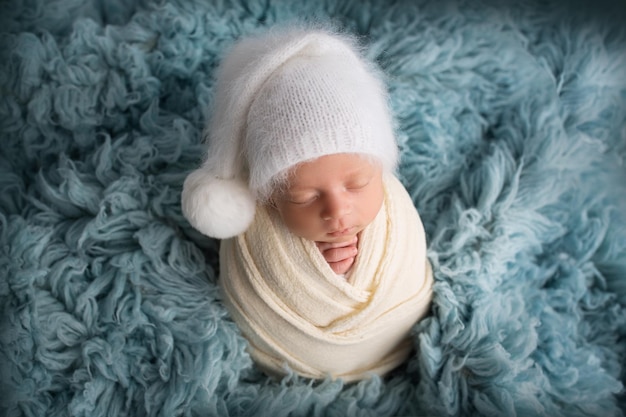 Slapende pasgeboren babyjongen in de eerste dagen van het leven in een witte zachte cocon met een gebreide wollen witte hoed op een blauwe achtergrond Studio macrofotografie portret van een pasgeboren Woman39s geluk