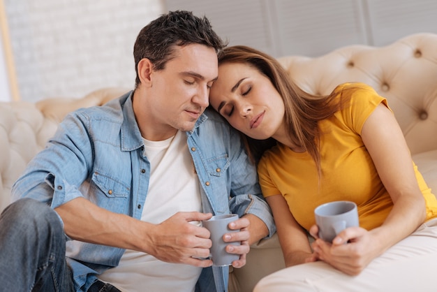 Slaperig stel. Vrij jong liefdevol paar dat samen met hun gesloten ogen zit en glimlacht terwijl twee mokken met lekkere warme thee worden vastgehouden