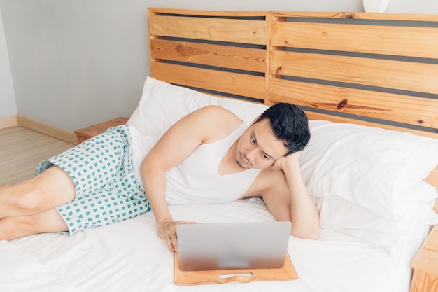 Slaperige man werkt met zijn laptop op zijn gezellige bed. Concept van saaie freelancer levensstijl.