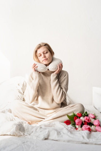 Slaperige vrouw zittend in bed met pyjama's en kussen over de nek, tulpenbloemen op het bed