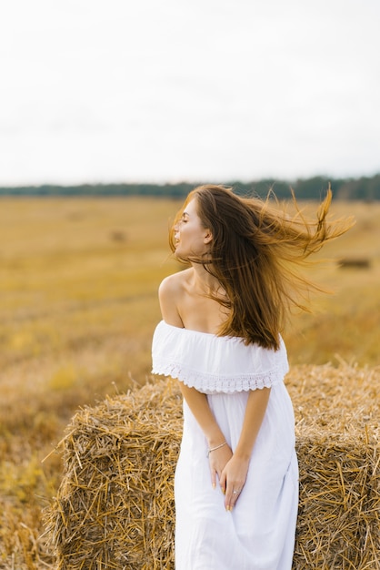 Slavic girl with long blond hair in a white dress standing in a field with bales of straw, her hair blowing in the wind