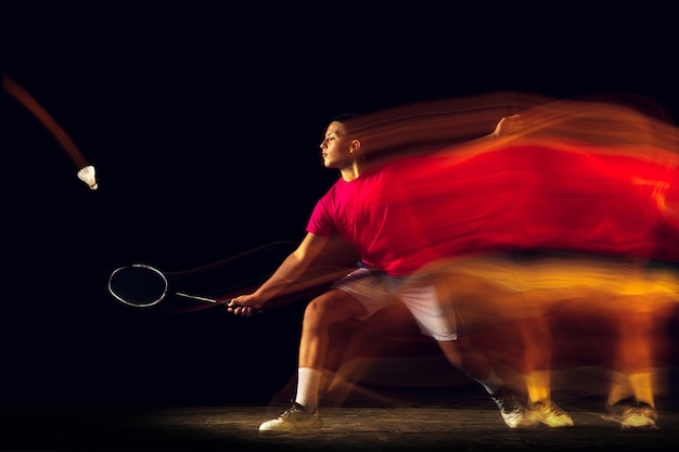 Slechts één stap om te winnen. Kleine jongen badminton spelen op zwarte achtergrond in gemengd licht. Jonge sportman in actie, beweging in het spel met de vuurschaduwen. Concept van sport, beweging, gezonde levensstijl.