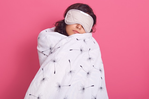 Sleep equipment concept. Portrait of brunette woman wrapped white blanket and wearing sleeping eye mask. Studio shot of young female isolated on pink, lady being ready to fall asleep.