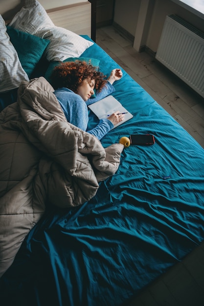 Спящая кавказская женщина с вьющимися волосами чувствует усталость после выполнения домашнего задания, держащего ручку и отдыха возле книги и мобильного телефона