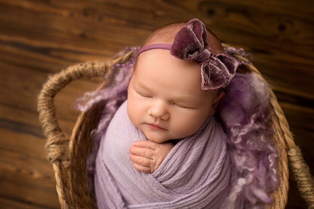 Photo sleeping newborn girl on a background of natural brown wood in a wicker basket a newborn baby with a lilacviolet bow on his head purple background inscription love beautiful portrait of a newborn