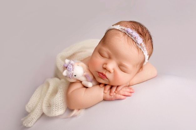 Sleeping newborn girl in the first days of life on a white\
background closeup studio portrait of a child a newborn baby is\
wrapped in a white knitted blanket with a white headband