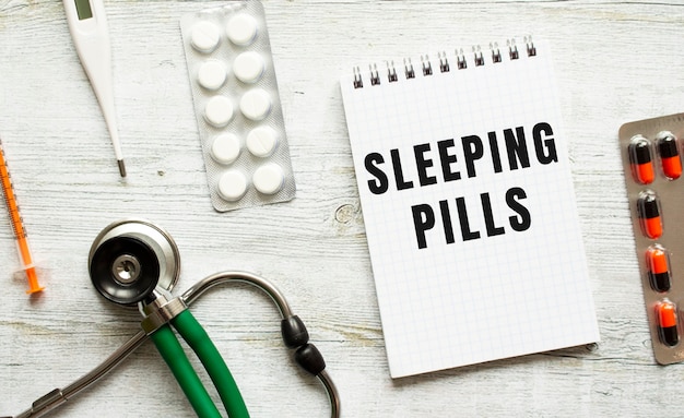 Sleeping pills is written in a notebook on a white table next to pills and a stethoscope