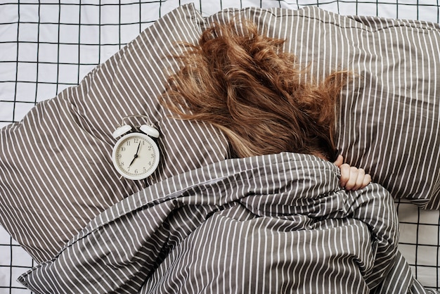 Sleeping woman covered under blanket in the bed and vintage alarm clock on pillow. Lazy morning ana wake up concept