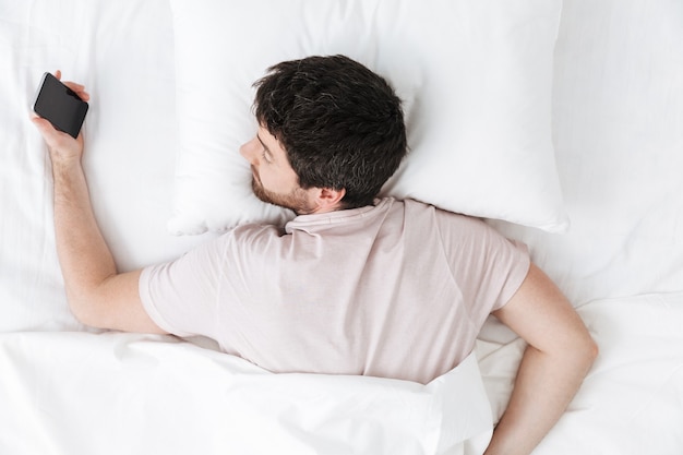 Спящий молодой человек утром под одеялом в постели лежит с помощью мобильного телефона