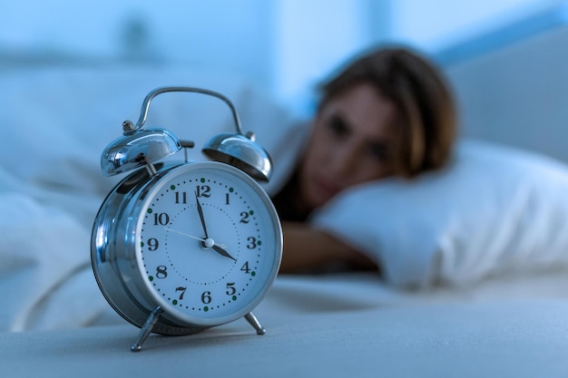 잠 못 이루고 절망적인 아름다운 백인 여성은 밤에 잠을 못 이루며 수면 장애 개념에서 불면증으로 고통받는 시계를 보며 좌절하고 걱정합니다.