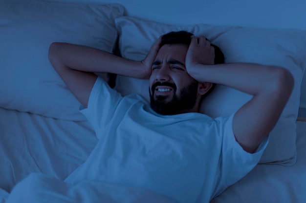 眠れないインド人男性が夜にベッドに横たわっている間に急性頭痛に苦しんでいます