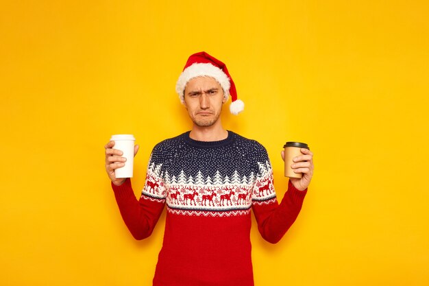 sleepless man in Christmas sweater Santa hat with paper cups of coffee in hands Space for text