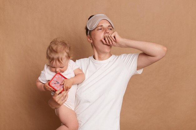 Sleepless woman wearing white t shirt and blindfold yawning and covering her mouth holding infant daughter kid holds red alarm clock has not enough sleep posing isolated over brown background