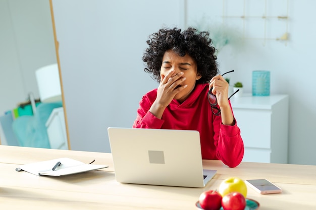 Sleepy african american woman sitting at table with laptop and yawning tired overworked girl lazy to