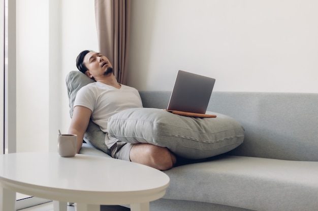 Photo sleepy asian man working on his laptop on the sofa.