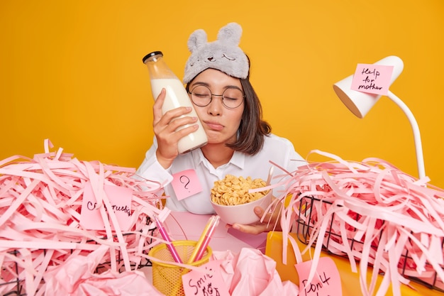 Sleepy asian woman had not enough sleep holds bottle of milk\
bowl with cornflakes spends time for course work poses at desktop\
wears sleepmask on forehead isolated over yellow studio wall