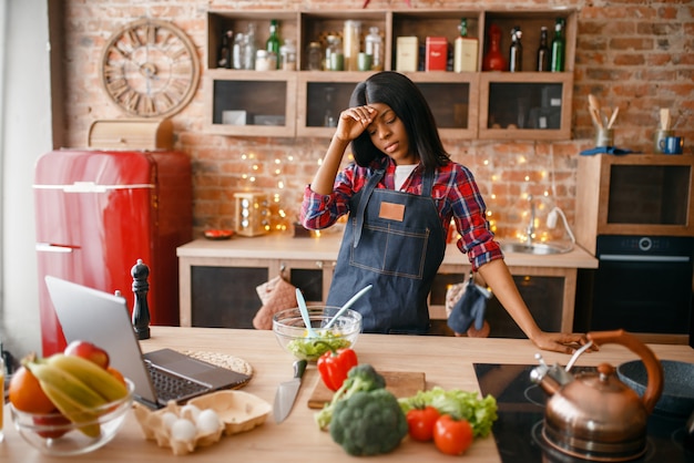 Foto donna nera sonnolenta in grembiule che cucina la colazione sana sulla cucina. persona di sesso femminile africana che prepara insalata di verdure a casa