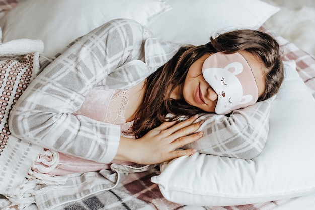 A sleepy brunette girl lies in bed with a blindfold in cute pajamas