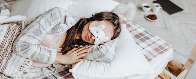 A sleepy brunette girl lies in bed with a blindfold waking up in the morning. Cozy home morning in cute pajamas. Have a good time, relax and dream. Healthy sleep.