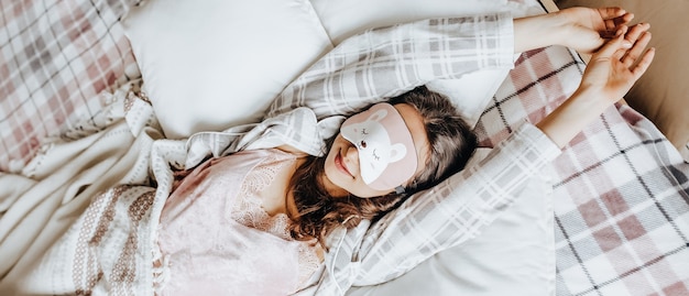 A sleepy brunette girl lies in bed with a blindfold waking up in the morning