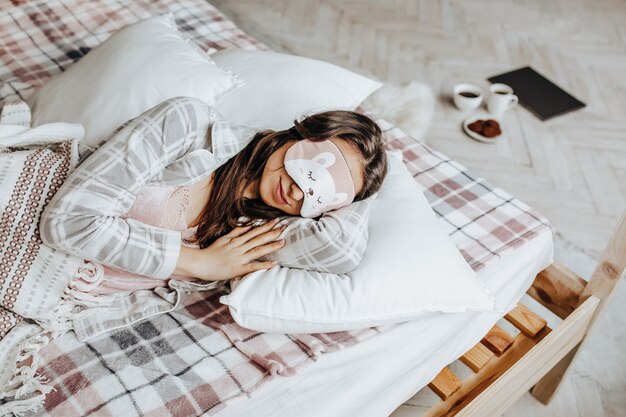 A sleepy brunette girl lies in bed with a blindfold waking up in the morning