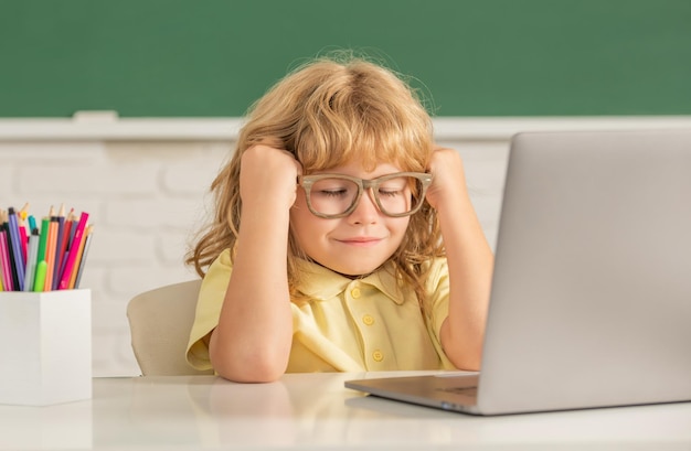Sleepy child boy in glasses study online in school classrrom with laptop education
