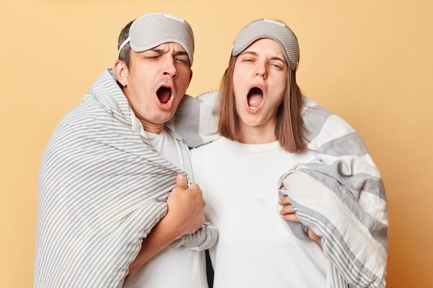 잠자는 커플: 담요로 감싸진 남녀, 베이지색 배경 위에 고립되어, 눈을 감고 서서, 입을 넓게 열고 는, 충분한 수면을 얻지 못한다.