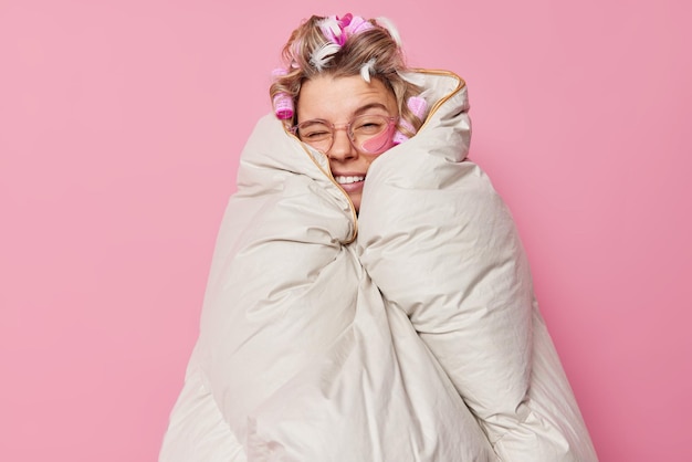Foto la sonnolenta donna europea strizza gli occhi avvolti in una coperta fa l'acconciatura con i bigodini indossa occhiali trasparenti isolati su sfondo rosa. risveglio mattutino e atmosfera domestica