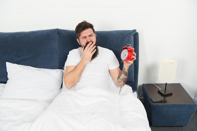 Una serata assonnata in camera da letto uomo barbuto che guarda l'orologio la sera pantaloni a vita bassa brutali sdraiati a letto in tarda serata impostazione della sveglia da sera a mattina