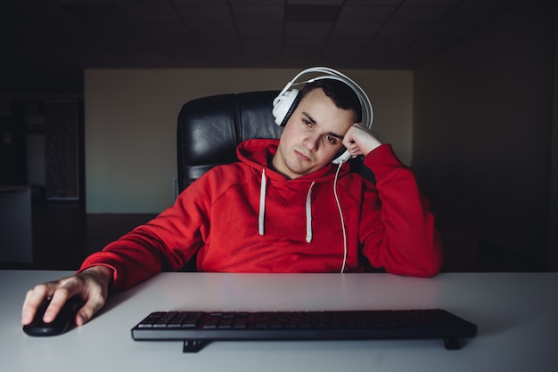 眠そうなゲーマーが自宅のコンピューターでビデオゲームをプレイしています。コンピューターの近くにヘッドフォンを持つ若者。