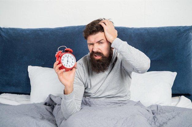 Sleepy guy holding alarm clock being in bed in morning oversleep