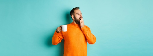 Sleepy handsome man drinking coffee and yawning standing in orange sweater against light blue person