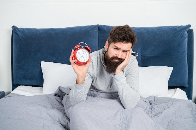 朝の時間にベッドにある目覚まし時計を保持している眠そうな男