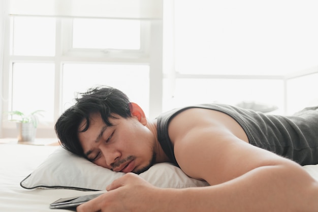 眠そうな男がベッドに横になってスマートフォンを使用しています。