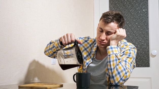 Sleepy man pours coffee while sitting at the table in the kitchen