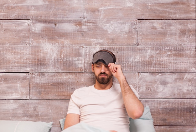 Sleepy man wearing pajamas in bed with sleep mask.