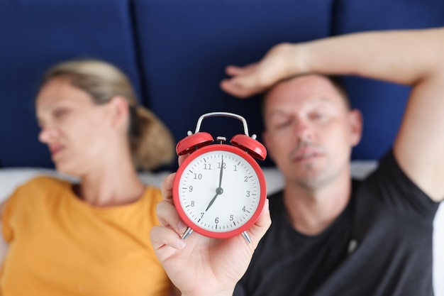 Сонный мужчина и женщина, лежа в постели и держащий красный будильник крупным планом