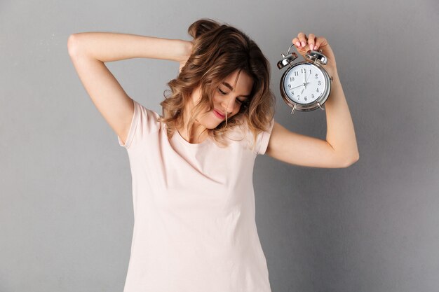 Sleepy smiling woman in t-shirt wake up while holding alarm clock with closed eyes over grey