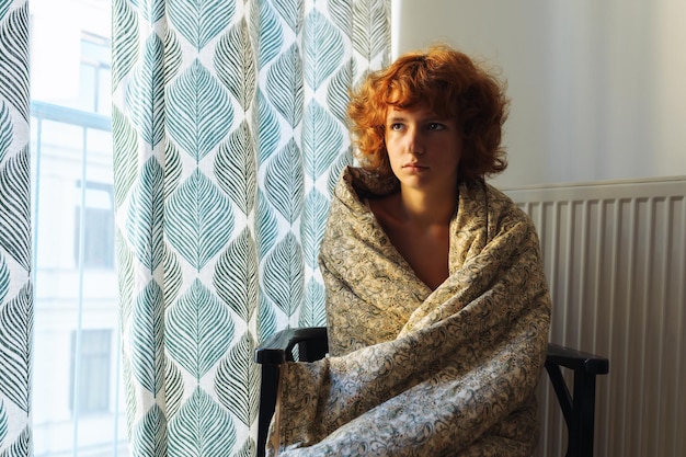 Sleepy teenage girl in blanket sitting on chair near radiator